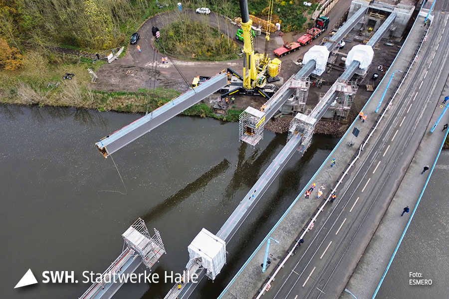 Eine Luftaufnahme. An einem großen gelben Kran hängt ein langes Stück Stahl direkt über dem Fluss, welcher von links nach rechts durch das Bild geht. Am rechten Bildrand ist die jetzige Elisabethbrücke zu sehen, auf de Personen stehen.